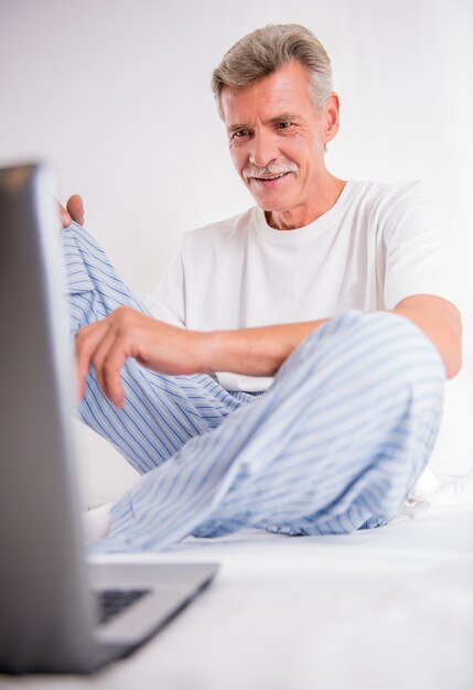 Alter Mann benutzt Laptop beim Sitzen im weißen Bett.