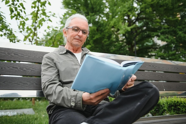 Alter Mann mit grauem Haar liest ein Buch auf einer Bank im Park Ruhen Sie sich im Park aus