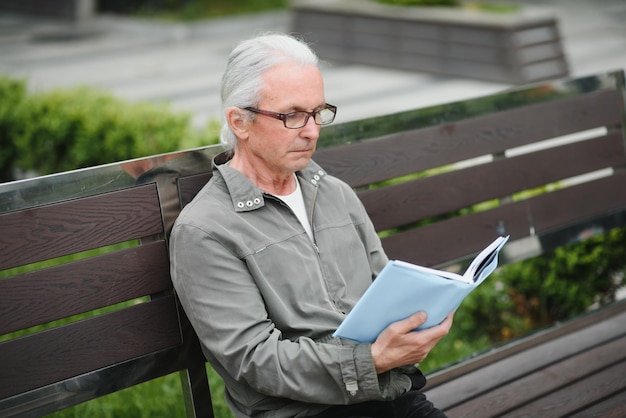 Alter Mann mit grauem Haar liest ein Buch auf einer Bank im Park Ruhen Sie sich im Park aus