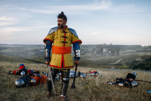 Alter mittelalterlicher Ritter in Rüstung sieht die Toten nach großer Schlacht an. Gepanzerter alter Krieger in der Rüstung, die auf dem Feld aufwirft