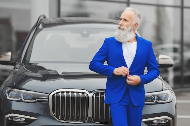 Foto alter styish mann, der nahe dem luxusauto steht. reifer reicher mann im anzug draußen in der nähe des autos.