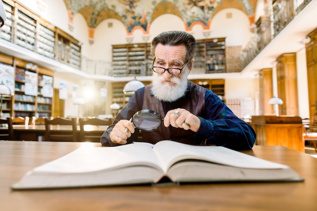 Alter Wissenschaftler, Bibliotheksarbeiter, liest ein Buch in einer Bibliothek und schaut durch die Lupe
