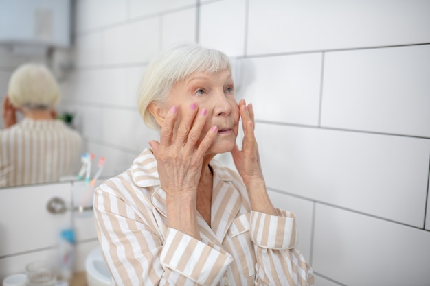 Altern. Grauhaarige Frau, die in den Spiegel im Badezimmer schaut und Wangen berührt