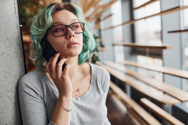 Alternatives Mädchen in Freizeitkleidung und mit grünen Haaren, das tagsüber drinnen steht und telefoniert.