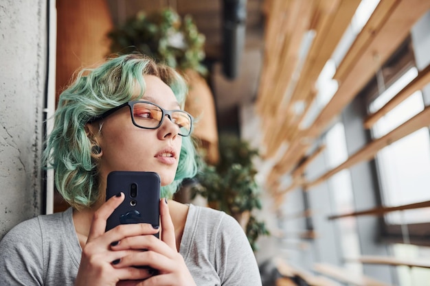 Foto alternatives mädchen mit brille und grünem haar, das tagsüber mit telefon in der hand drinnen sitzt