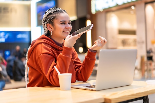 Alternatives Mädchen mit weißen Zöpfen, das einen Computer in einem Einkaufszentrum benutzt und lächelt und eine Audionachricht macht