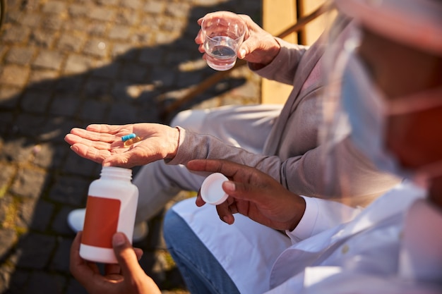 Alternde Person, die auf einer Bank mit blauen und orangefarbenen Pillen in ihrer Handfläche sitzt, während sie ein Glas Wasser hält