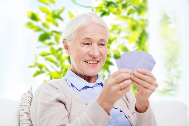 Alters-, Ruhestands- und Personenkonzept - glücklich lächelnde ältere Frau, die zu Hause Karten über Fenster und grünem Naturhintergrund spielt