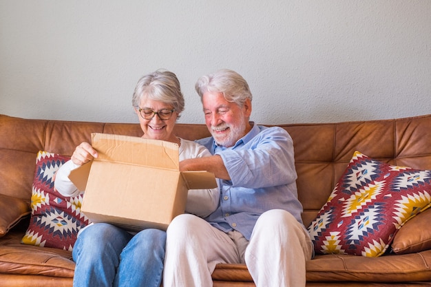 Altes Ehepaar, das zu Hause Lieferkarton auspackt. Glückliches älteres Paar, das Karton beim Sitzen auf dem Sofa im Wohnzimmer zu Hause betrachtet. Älteres Ehepaar, das zu Hause gelieferte Sachen auscheckt.