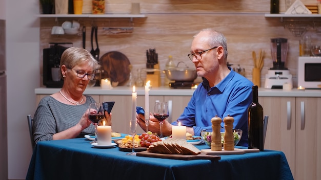 Altes Ehepaar im Ruhestand mit Smartphones beim romantischen Abendessen. In der Küche am Tisch sitzen, stöbern, suchen, telefonieren, Internet nutzen, im Esszimmer Jubiläum feiern.