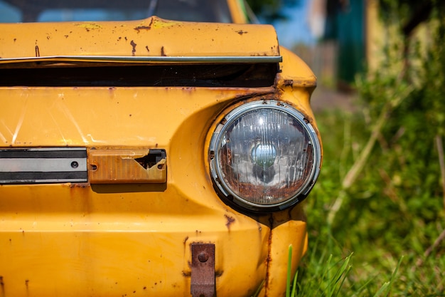 Altes gelbes Autowrack im Vintage-Stil. Verlassenes rostiges gelbes Auto. Nahaufnahme der Scheinwerfer der Vorderansicht eines rostigen, kaputten, verlassenen Autos in der Nähe des Hauses. Konzept des verlassenen Gebrauchtwagens.