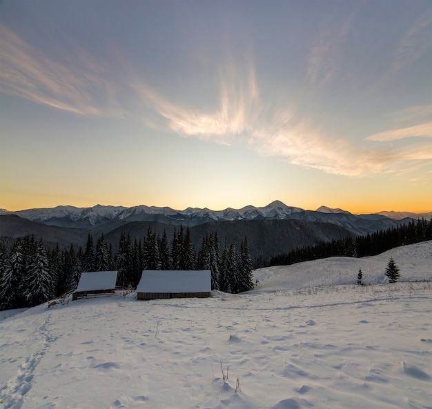 Altes Holzhaus, Hütte und Scheune im tiefen Schnee auf Gebirgstal, Fichtenwald, waldige Hügel auf klarem blauem Himmel am Sonnenaufgangkopien-Raumhintergrund. Gebirgswinter-Panoramalandschaft.