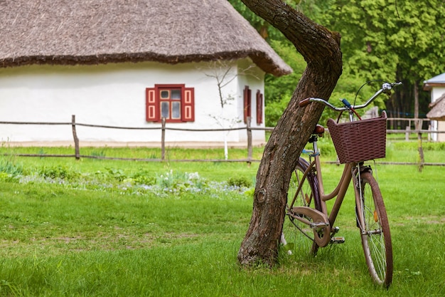 Altes ukrainisches Haus mit einem Strohdach und einem Fahrrad mit einem Korb in der Nähe des Baumes