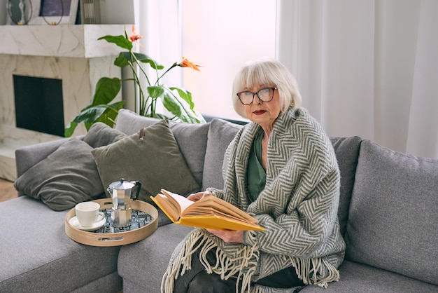 altmodische ältere Frau, die auf der Couch sitzt und zu Hause ein Buch liest