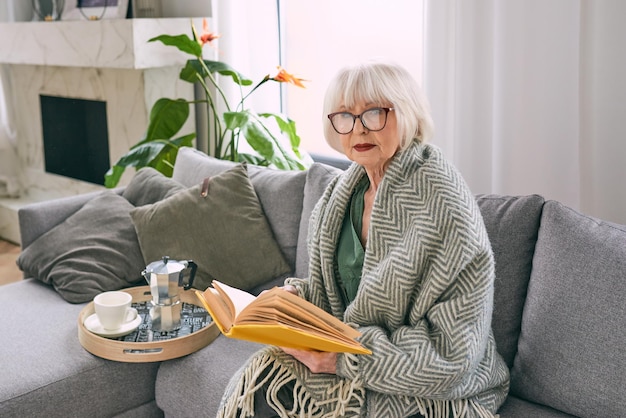 altmodische ältere Frau, die auf der Couch sitzt und zu Hause ein Buch liest