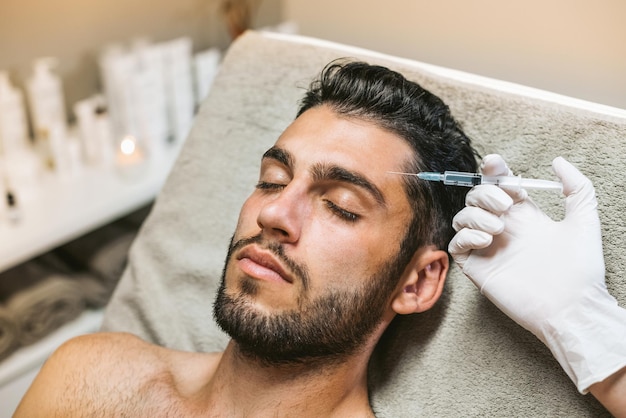 Foto un alto ángulo de un cliente masculino barbudo cerrando los ojos mientras un esteticista inyecta botox en la frente con una jeringa en un salón de belleza