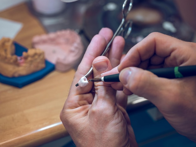 Alto ángulo de cultivo anónimo especialista dental masculino haciendo dientes cerámicos artificiales para dentaduras postizas mientras trabaja con herramientas profesionales en un laboratorio moderno