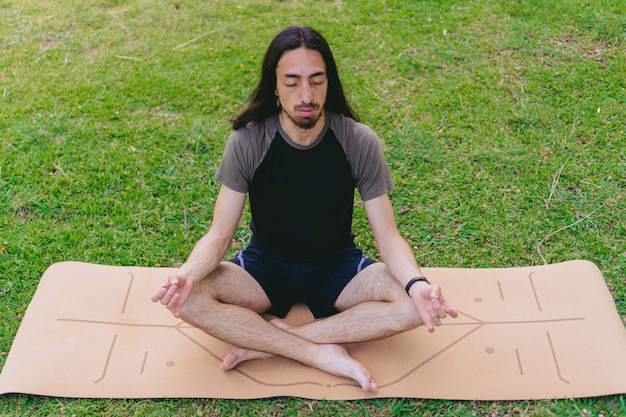 Alto ângulo de um jovem hippie hispânico e latino meditando com os olhos fechados enquanto faz a pose de lótus em um tapete de ioga em um pasto
