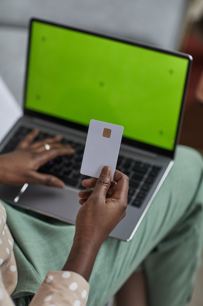 Alto ângulo vertical close-up de uma jovem afro-americana usando o laptop com tela verde e segurando o cartão de crédito, copie o espaço