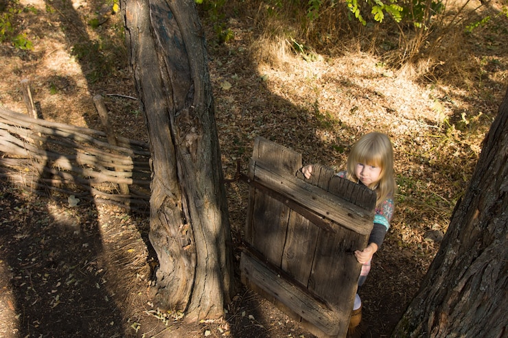 Foto un alto ángulo de vista de una joven rubia jugando con una vieja puerta de madera entre dos árboles en un jardín mirando hacia la cámara