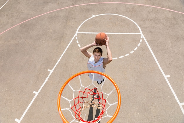 Un alto ángulo de vista desde el tablero del joven Atlético haciendo tiro en salto en la red en la cancha de baloncesto al aire libre