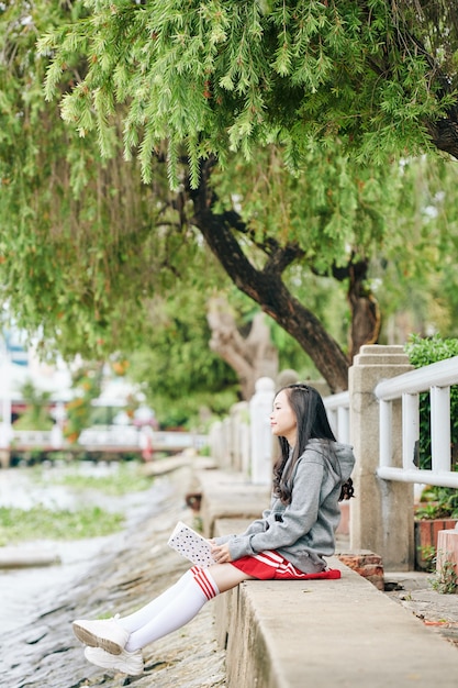 Alumna coreana pensativa con libro en manos sentado en la orilla del río y mirando el agua