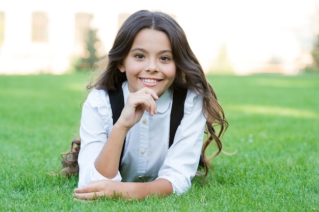 Alumno sonriente feliz. Niño lindo de la muchacha que pone la hierba verde. Niño feliz relajándose al aire libre. Uniforme escolar de niña disfruta relajarse. Colegiala alegre. Relájese en el patio de la escuela. Buen tiempo. Receso escolar para descansar.