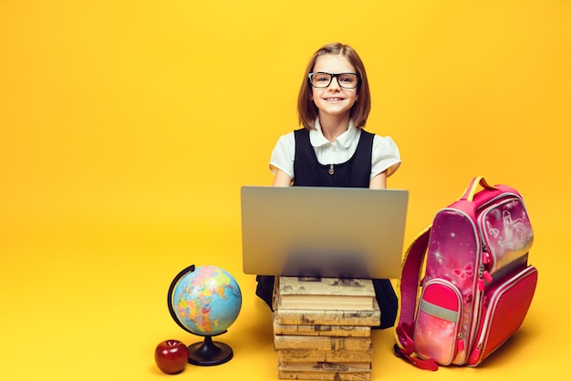 Alumno sonriente se sienta detrás de una pila de libros y una computadora portátil con globo mira la educación de los niños de la cámara