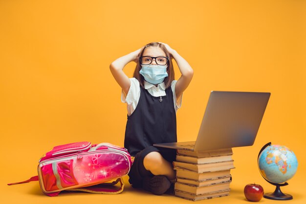 Alumno sorprendido en máscara médica se sienta detrás de una pila de libros y educación infantil portátil en la pandemia