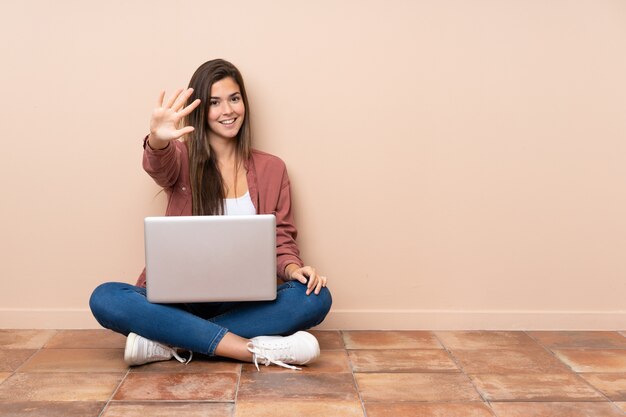 Aluna adolescente sentado no chão com um laptop contando cinco com os dedos