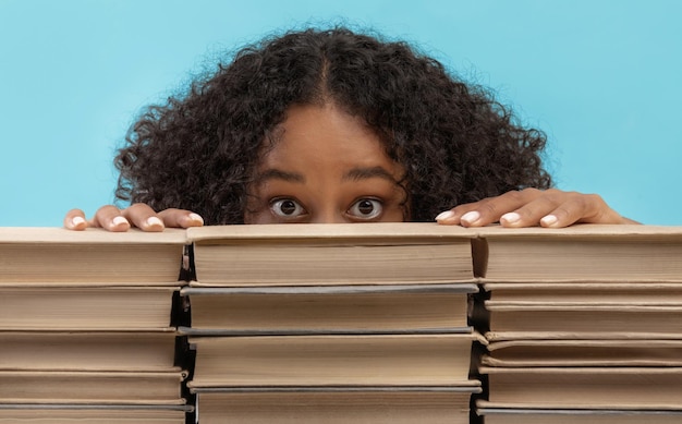 Foto aluna afro-americana assustada se escondendo atrás de pilhas de livros com medo do exame universitário em