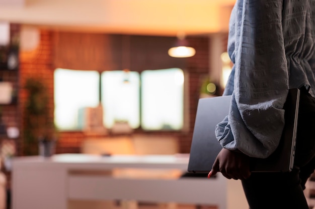 Aluna afro-americana carregando laptop na sala de casa com interior moderno. Jovem empresária em roupas casuais segurando computador portátil, vista lateral com fundo desfocado e espaço de cópia