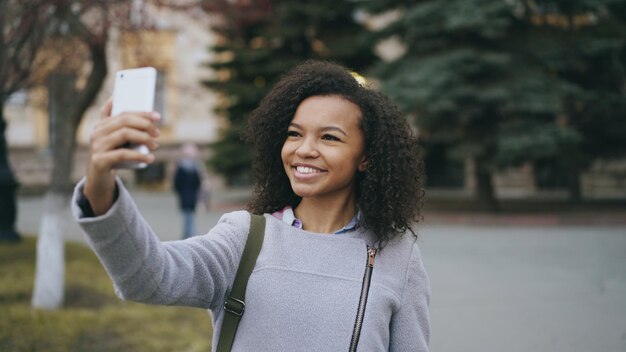 Aluna alegre de raça mista falando em videochamada com smartphone perto da universidade