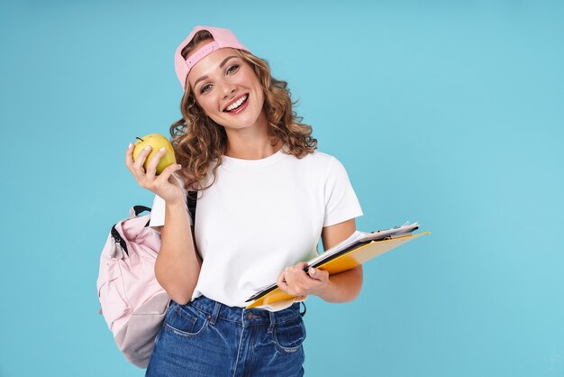 Aluna alegre e bonita carregando uma mochila e segurando livros didáticos e uma maçã em pé isolado no azul