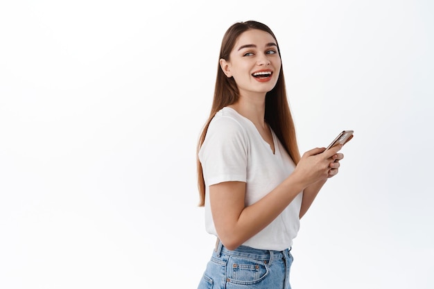 Aluna alegre em seu telefone, parece feliz atrás do ombro no texto promocional do espaço de cópia, usando o aplicativo de bate-papo do smartphone, mídia social, em pé sobre um fundo branco