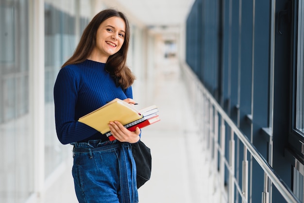 Foto aluna alegre morena com mochila preta segurando livros em um prédio moderno