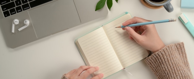 Aluna, anotando no caderno em branco ao fazer a tarefa com o laptop na mesa de estudo