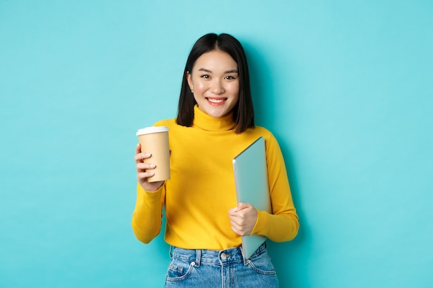 Aluna asiática elegante com uma xícara de café em pé sobre fundo azul, segurando o laptop na mão, sorrindo para a câmera, em pé sobre fundo azul.