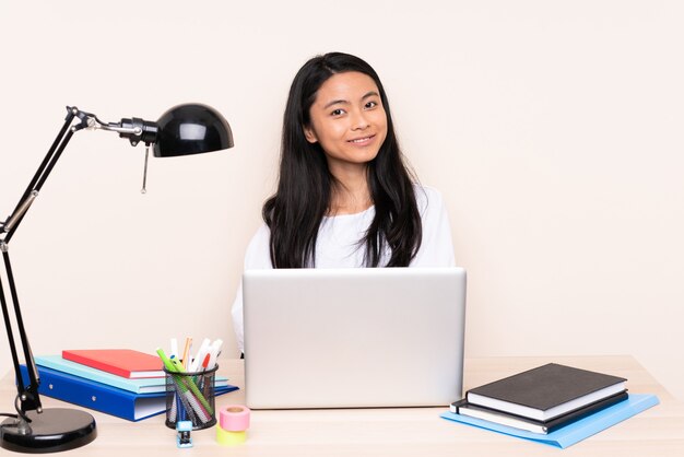 Aluna asiática em um local de trabalho com um laptop isolado em bege com os braços cruzados e feliz