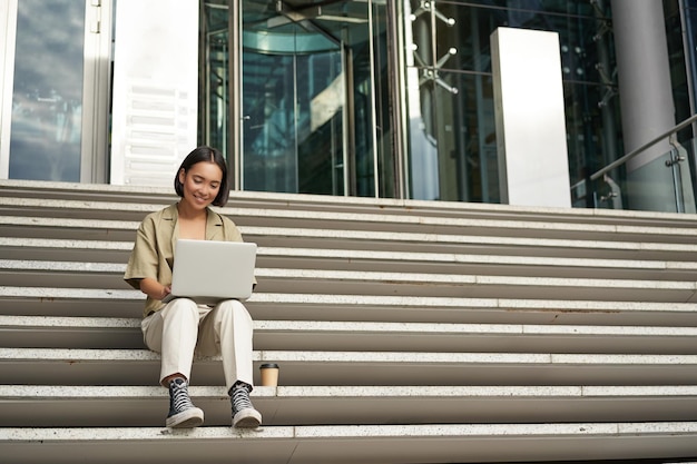 Aluna asiática senta-se em escadas perto de tipos de campus no laptop faz sua lição de casa ao ar livre