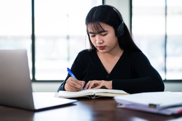 Aluna asiática sorridente usando fones de ouvido sem fio escreva no notebook para estudar idiomas online, assistir e ouvir o palestrante, webinar via videochamada e-learning em casa, educação à distância