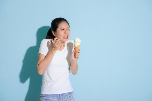 aluna atraente bonita depois de comer um delicioso sorvete sentindo os dentes sensíveis dolorosos desconfortáveis e fazendo expressão de tristeza no fundo da parede azul.