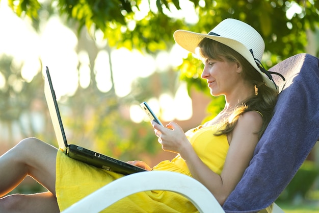 Aluna com vestido de verão amarelo, descansando no gramado verde no parque de verão, estudando em mensagens de texto de computador portátil no celular móvel.