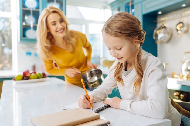 Aluna de cabelo louro muito alegre, sorrindo e escrevendo em seu caderno, enquanto sua mãe está perto dela com uma panela