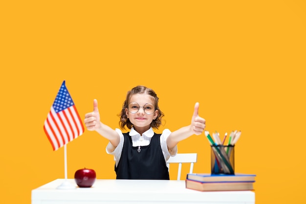Aluna de colegial caucasiana feliz sentada na mesa gesticulando com o polegar para cima, na aula de inglês, bandeira dos EUA