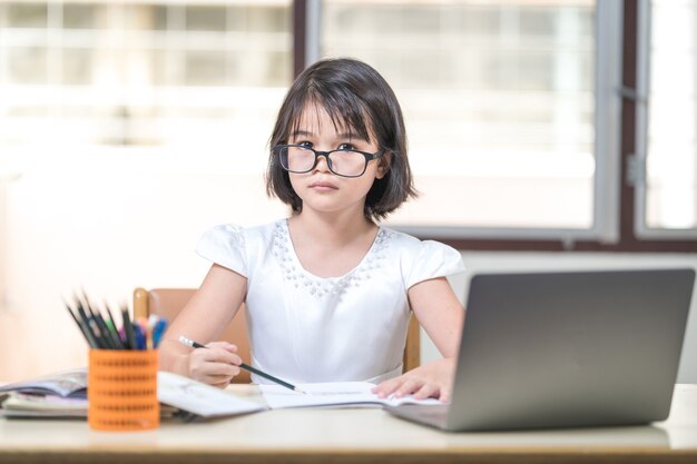 Aluna de crianças asiáticas com óculos, estudando online, fazendo lição de casa no laptop em casa. Conceito de educação Foto de stock