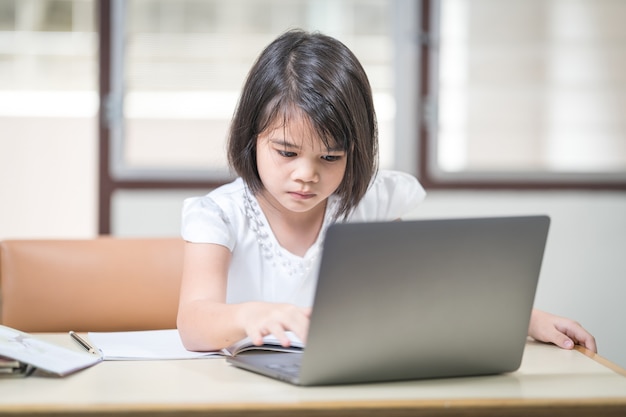 Aluna de crianças asiáticas escrevendo no caderno enquanto estudava online através do laptop enquanto estava em casa. Conceito de educação Foto de stock