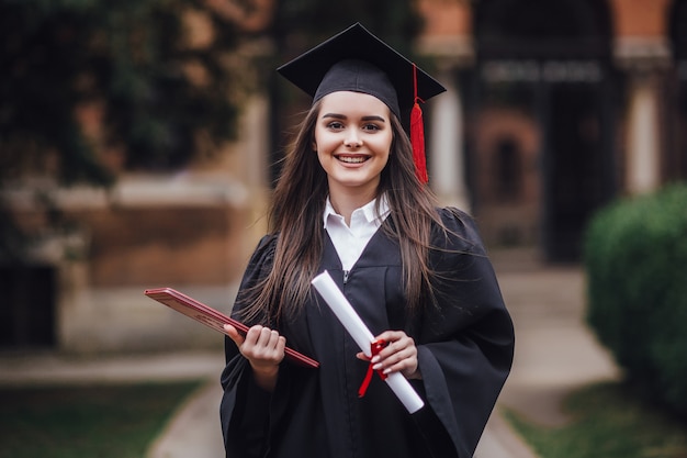 Foto aluna de graduação está de pé no salão da universidade em manto, sorrindo com diploma nas mãos.