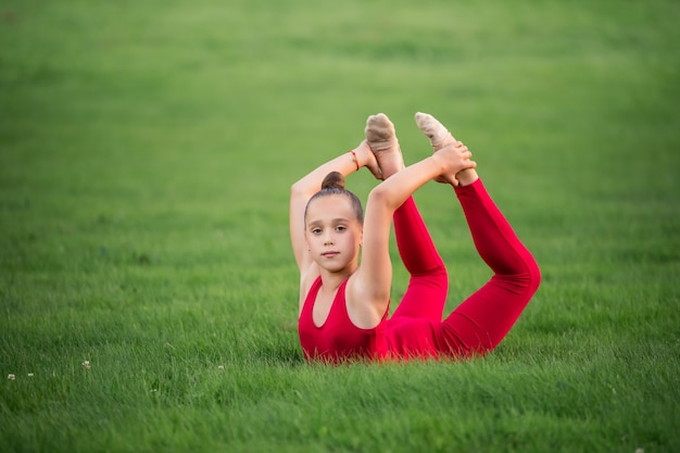 Aluna de macacão vermelho brilhante pratica ginástica na grama, fazendo exercícios de alongamento