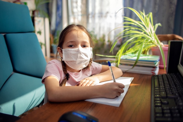 Aluna de menina em uma máscara médica faz lição de casa na frente de um computador